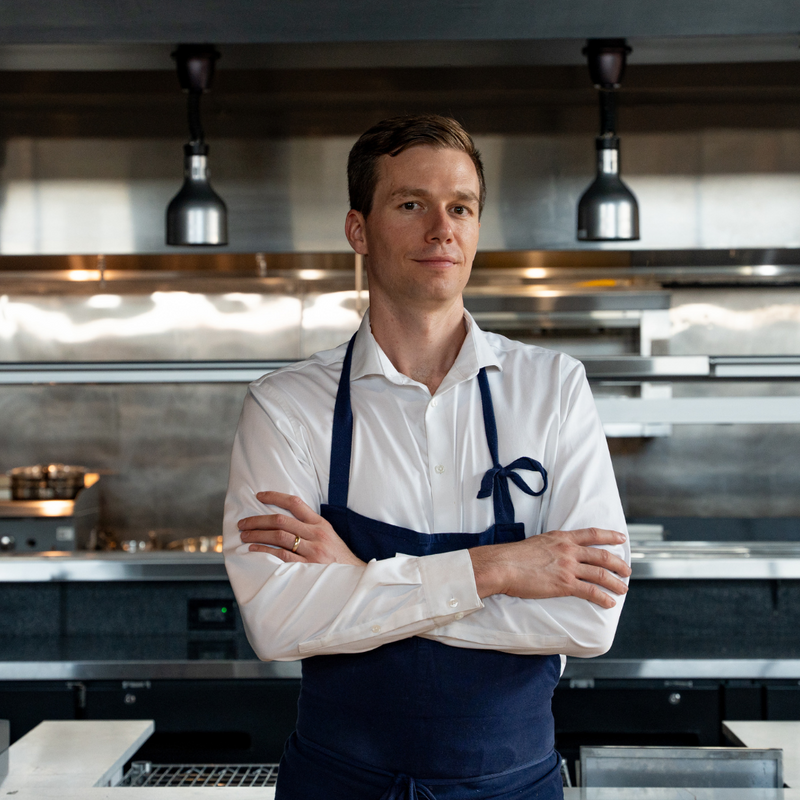 Chef Tyler Vorce in a white dress shirt and navy apron standing with arms crossed in a professional kitchen setting.